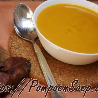 Kom romige pompoensoep zonder room, glad gepureerd en mooi oranje, geserveerd met lepel naast herfstbladeren.